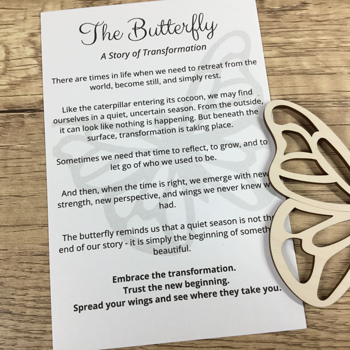 A printed card titled "The Butterfly: A Story of Transformation" with a poem about butterfly metamorphosis, placed on a wooden surface next to a light natural wood butterfly-shaped ornament.