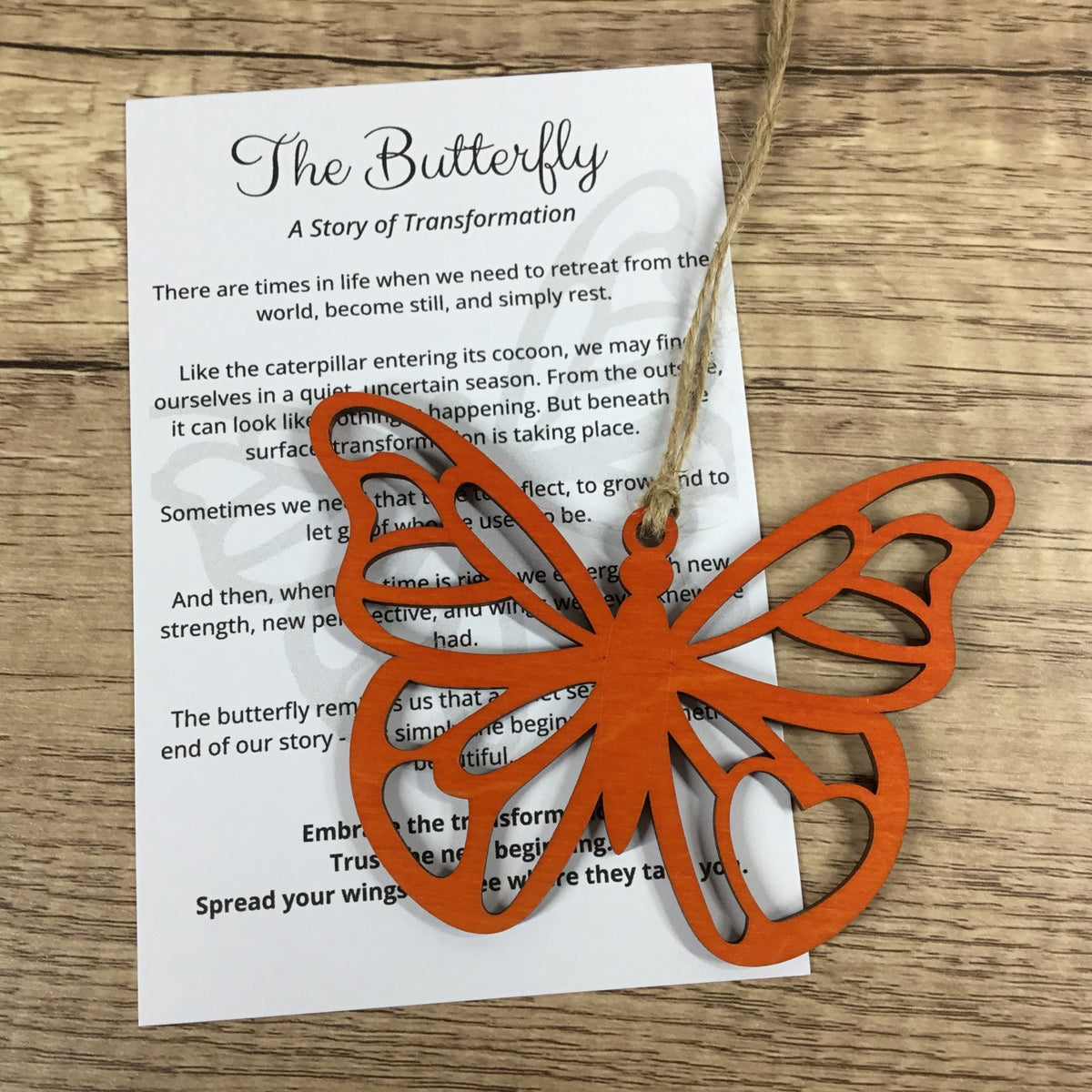 An orange laser-cut butterfly ornament hangs from a string over a printed white card titled 'The Butterfly: A Story of Transformation' on a wooden surface.