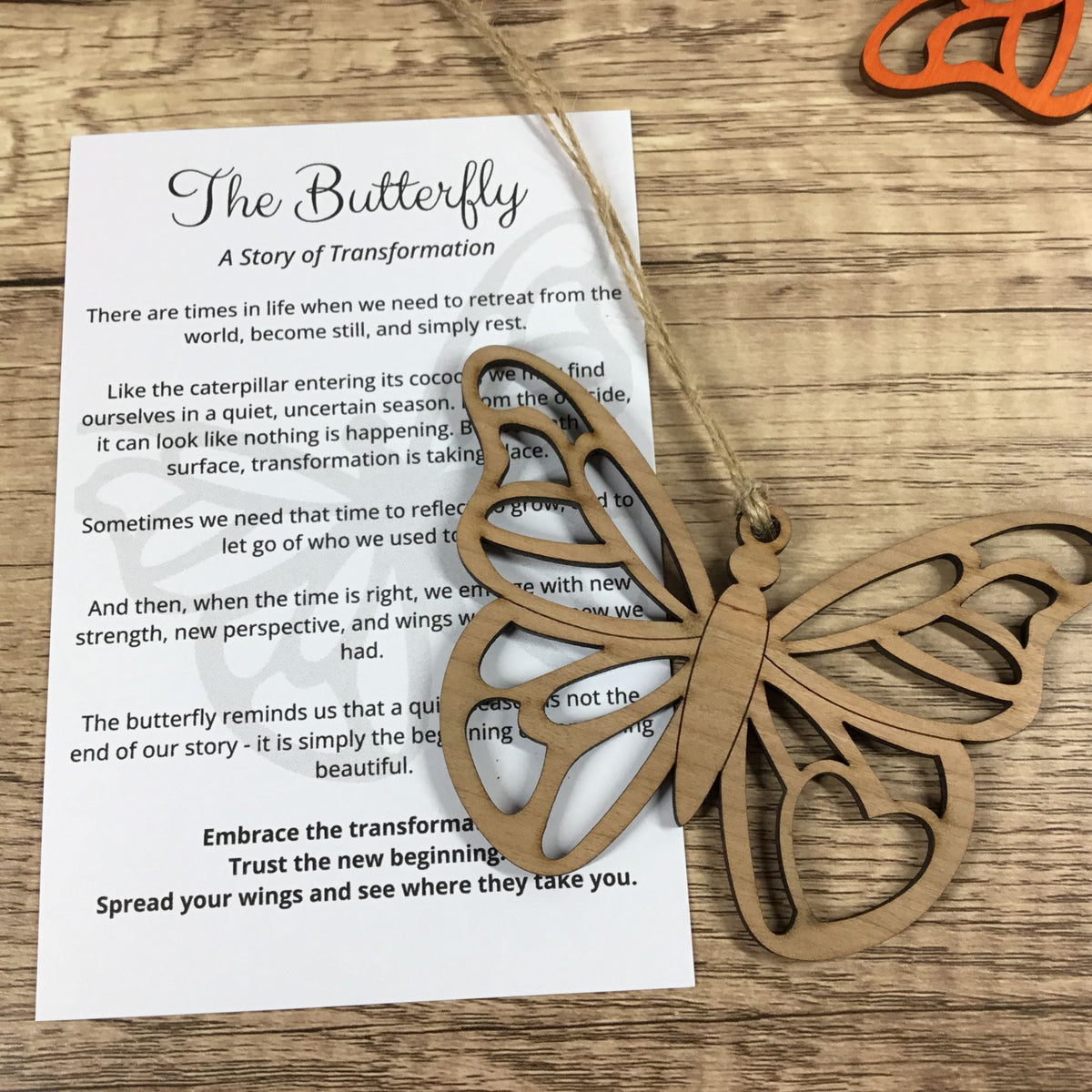 A laser-cut wooden butterfly ornament hanging over a printed card titled "The Butterfly: A Story of Transformation" on a wood surface, with an orange-handled tool partially visible in the corner.