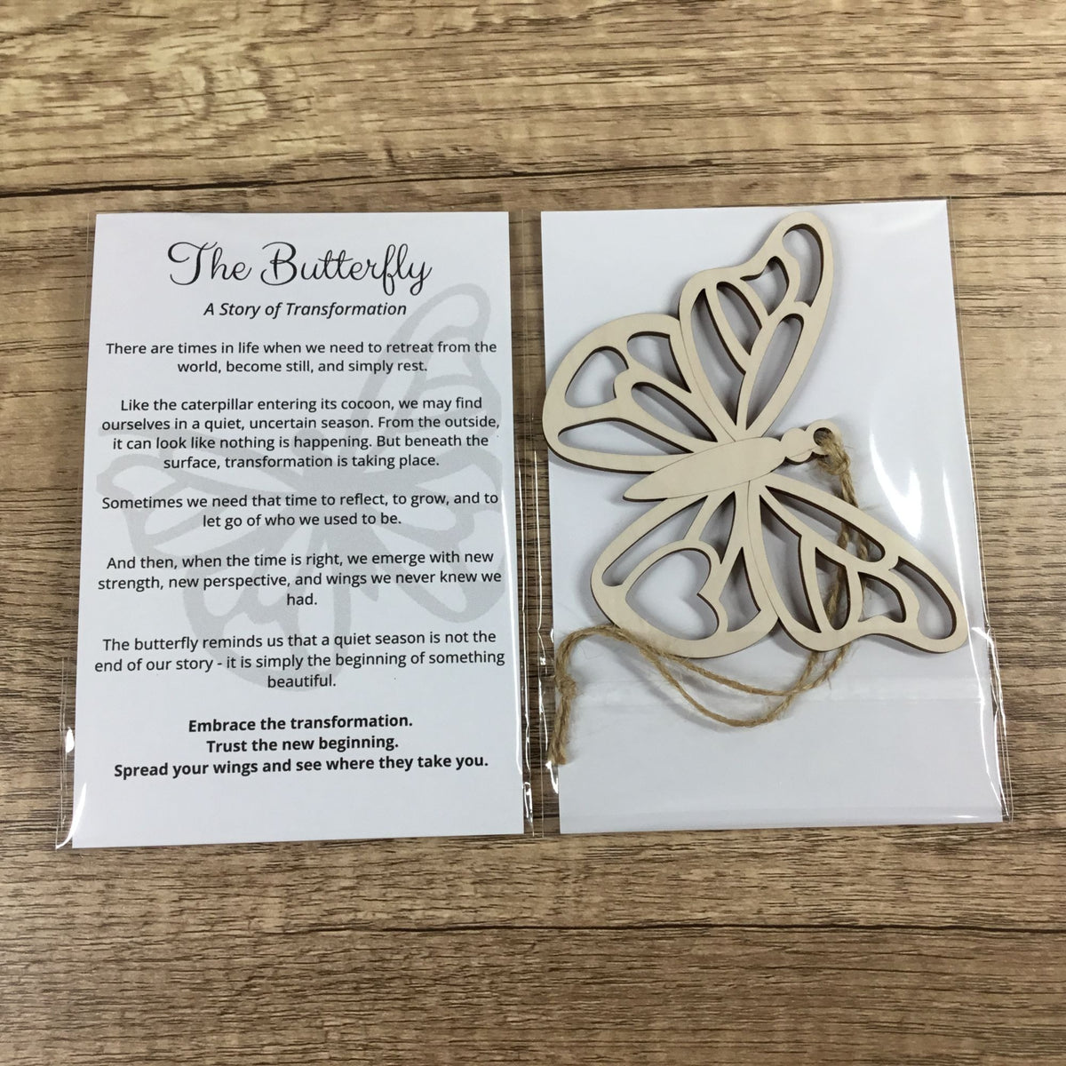 A wooden butterfly-shaped ornament in a clear plastic sleeve next to its printed backing card titled 'The Butterfly: A Story of Transformation,' placed on a wood surface.