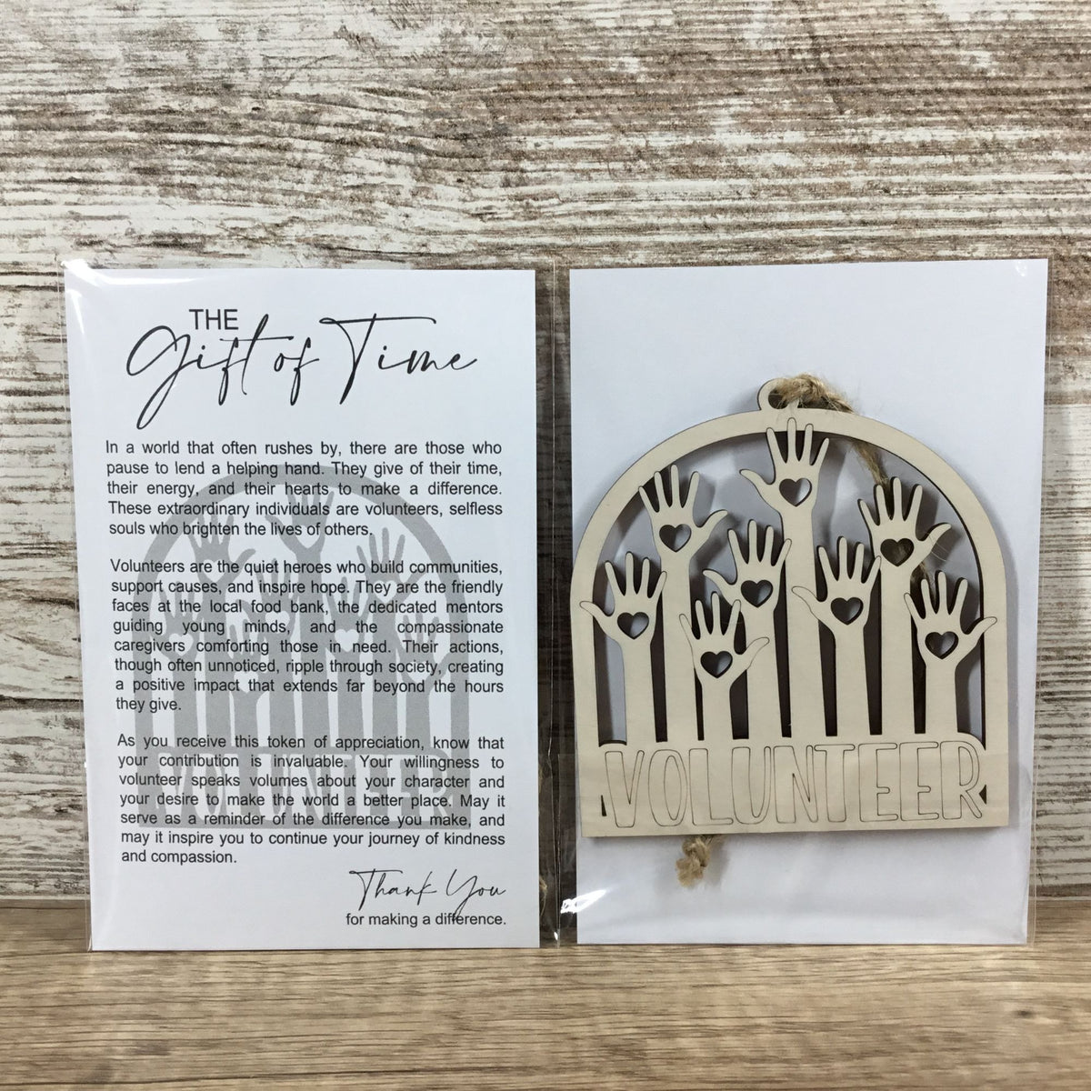 A packaged wooden volunteering ornament on a rustic wood background, shown next to its printed message card titled 'The Gift of Time'. The wooden plaque features handprints and the word 'VOLUNTEER'.