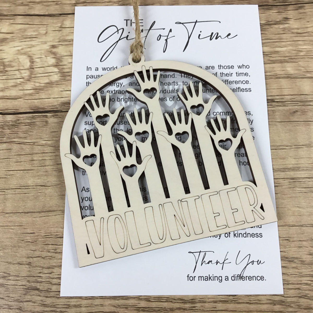 A laser-cut wooden arch-shaped hanging ornament with hand-shaped cutouts and the word “VOLUNTEER” cut into the bottom, displayed over a printed card on a wood surface.