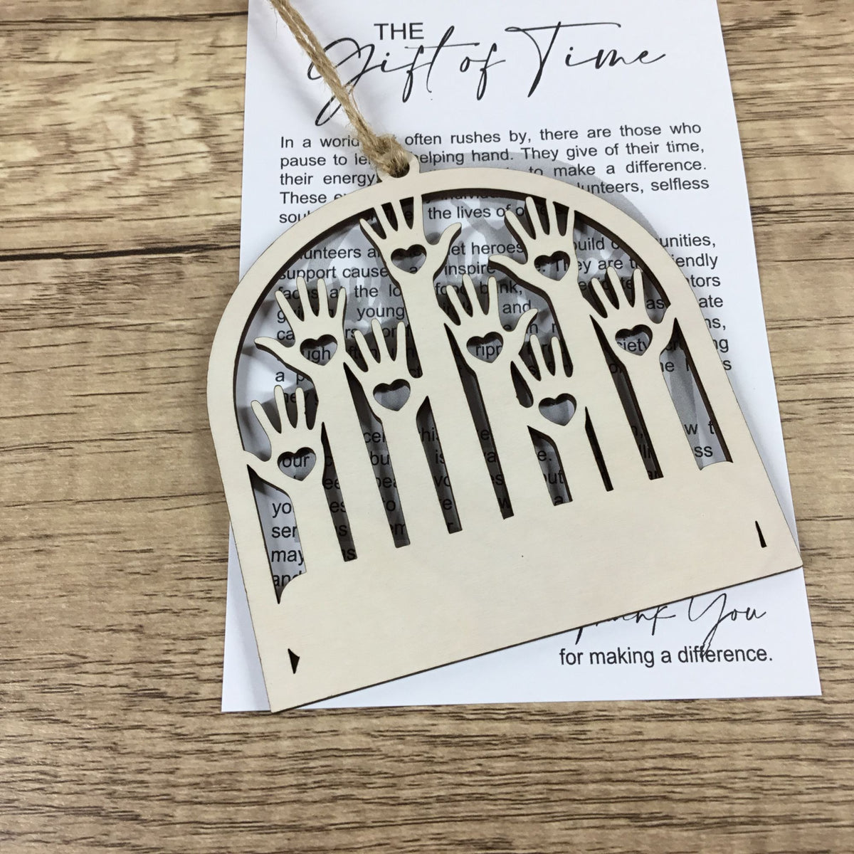 A light natural wood keepsake ornament with handprints and heart cutouts, hanging from a twine string over a printed card on a wooden surface.