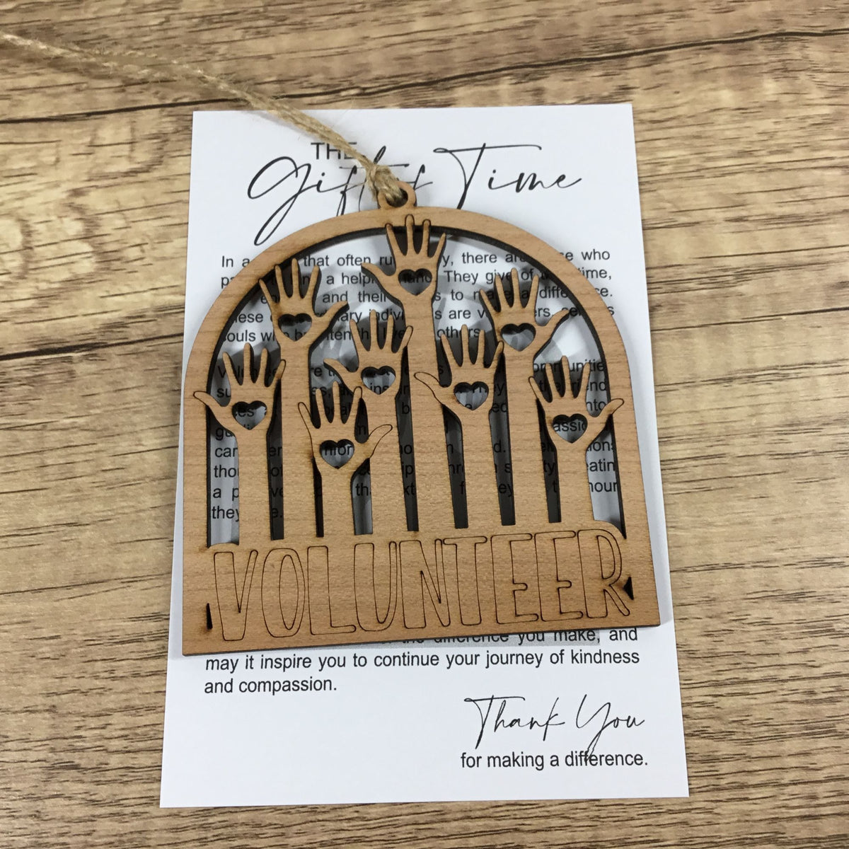 A wooden hanging ornament with laser-cut handprints and the word 'VOLUNTEER' on the front, displayed on top of a printed card over a wood surface.
