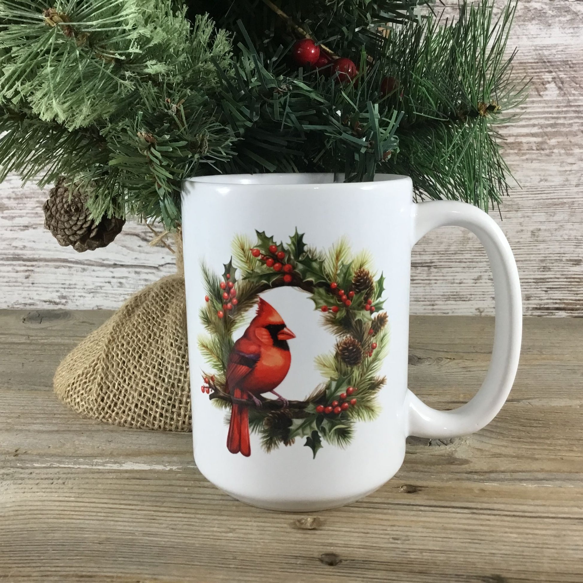 White mug with a red cardinal and holly design on a wooden surface with a Christmas tree in the background.