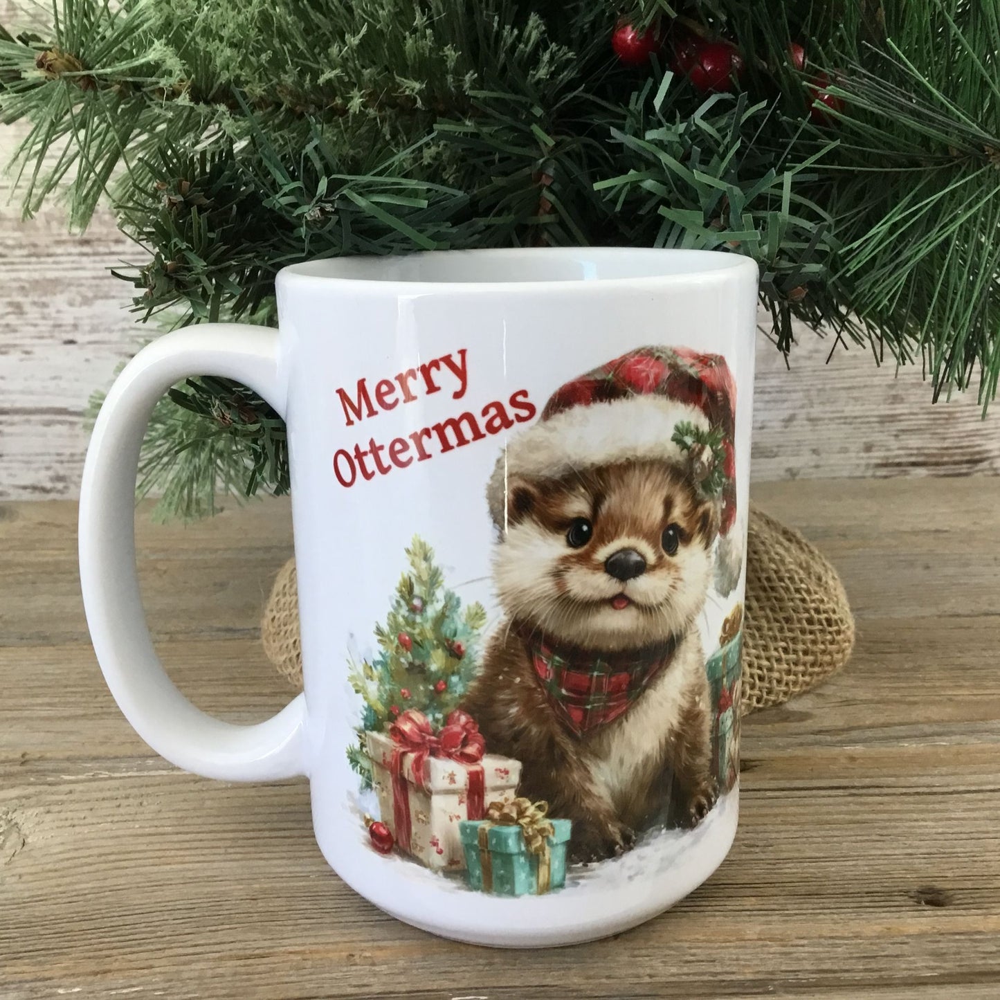 Mug with otter illustration and 'Merry Ottermas' text, placed on a wooden surface with Christmas decorations.