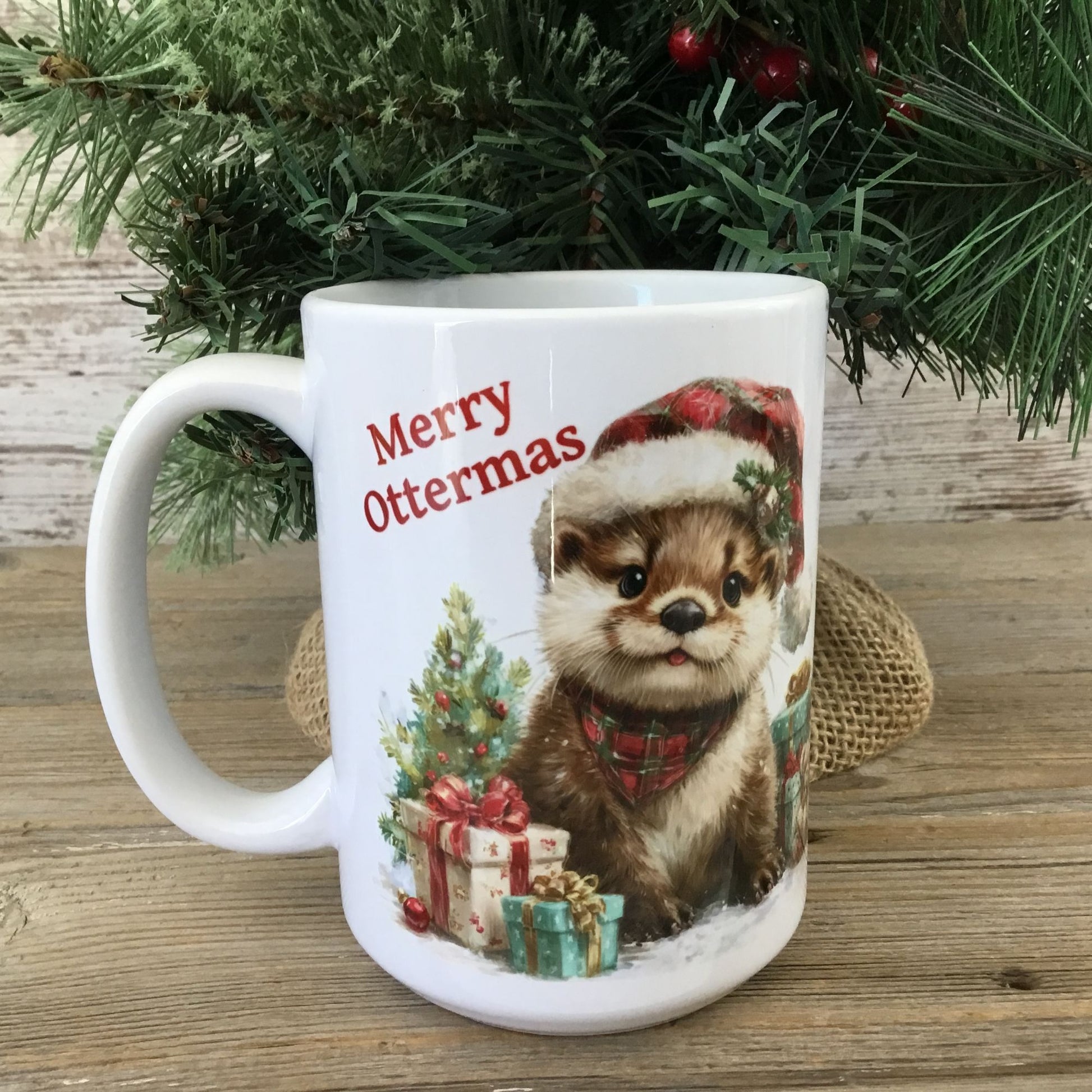 Mug with otter illustration and 'Merry Ottermas' text, placed on a wooden surface with Christmas decorations.