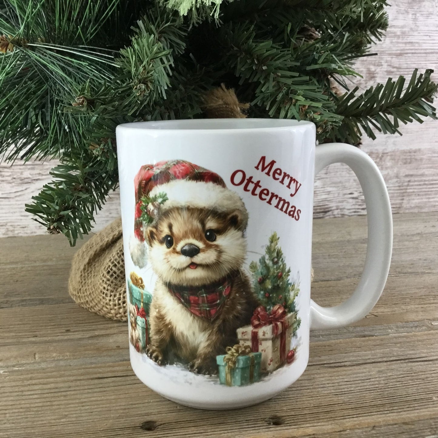 White mug with a Christmas otter design and 'Merry Ottermas' text, placed under a decorated tree.